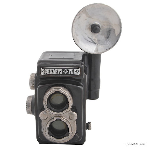  Ceramic schnapps holder or decanter, entitled “Schnapps-O-Flex”, and in the form of a camera, the flash bulb is the stopper, American, circa 1980s. Height: 8-1/2″ Width: 3-1/4″